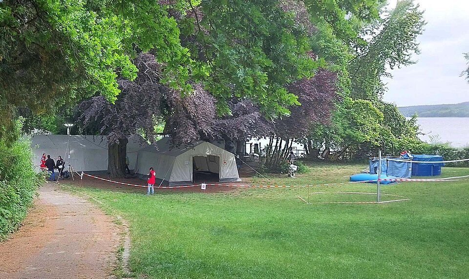 Auf einer Wiese am Wasser unter Bäumen stehen Zelte und eine mobile Trinkwasseranlage. Menschen mit und ohne Rotkreuzwesten stehen auf der Wiese
