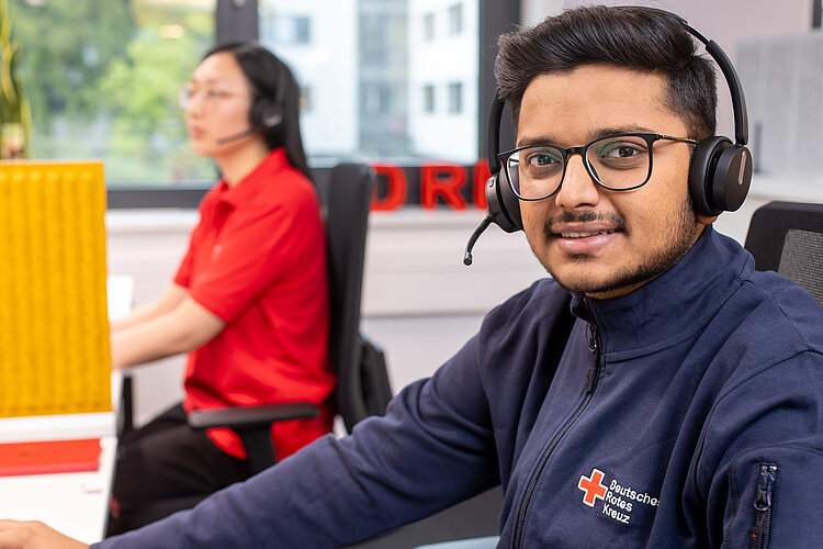 DRK-Mitarbeiter_mit_Headset Ein junger Mann mit Headset und blauem Shirt mit DRK-Abzeichen lächelt den Betrachter an. Im Hintergrund sitzt eine Frau mit Headset.