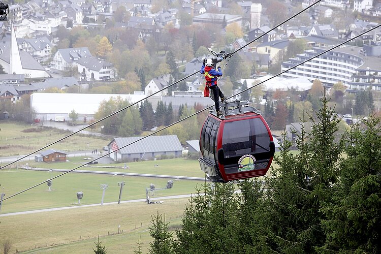 Ein Bergretter steht auf dem Dach einer roten Gondelbahn und sichert sich an den Seilen, während unter ihm dichte Tannen und im Hintergrund eine Ortschaft zu sehen sind. Die Szene zeigt eindrücklich die Arbeit der Bergrettung bei Einsätzen in luftiger Höhe.