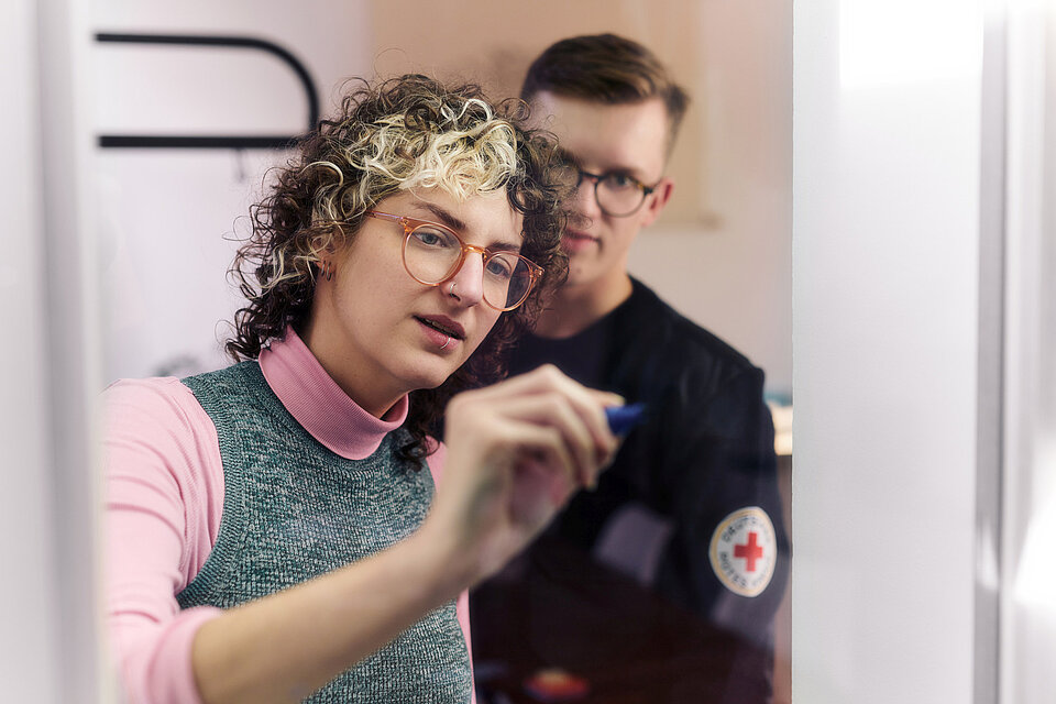DRK-Mitarbeiterin Mitarbeiterin des DRK schreibt mit einem Filzstift auf Glas, daneben steht ein Mitarbeiter mit DRK-Abzeichen