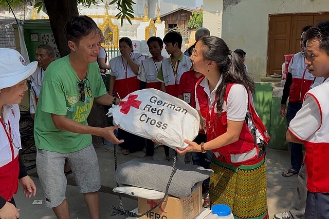 Auf dem Bild sieht man eine Szene nach dem Erdbeben 2025 in Myanmar: Eine Mitarbeiterin des Deutschen Roten Kreuzes überreicht einen Sack voller Hilfsgüter an Mitarbeiterinnen und Mitarbeiter des Myanmarischen Roten Kreuzes. Auf dem Sack steht geschrieben "German Red Cross". Die Beteiligten schauen sich gegenseitig freudig an. Die Mitarbeiterinnen und Mitarbeiter des Myanmarischen Roten Kreuzes tragen Westen ihrer Hilfsorganisation, während die Mitarbeiterin des Deutschen Roten Kreuzes neutral gekleidet ist.