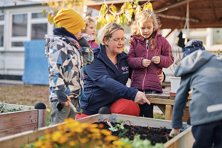 Vier kleine Kinder stehen an einem Hochbeet und eine Erzieherin im blauen Shirt mit Rotkreuz-Logo zeigt auf etwas