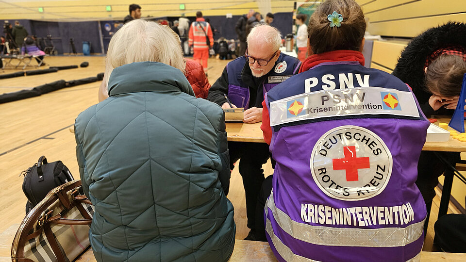 In einer Turnhalle sitzen Menschen an einem Tisch. PSNV-Einsatzkräfte des DRK in violetten Westen nehmen Daten auf.